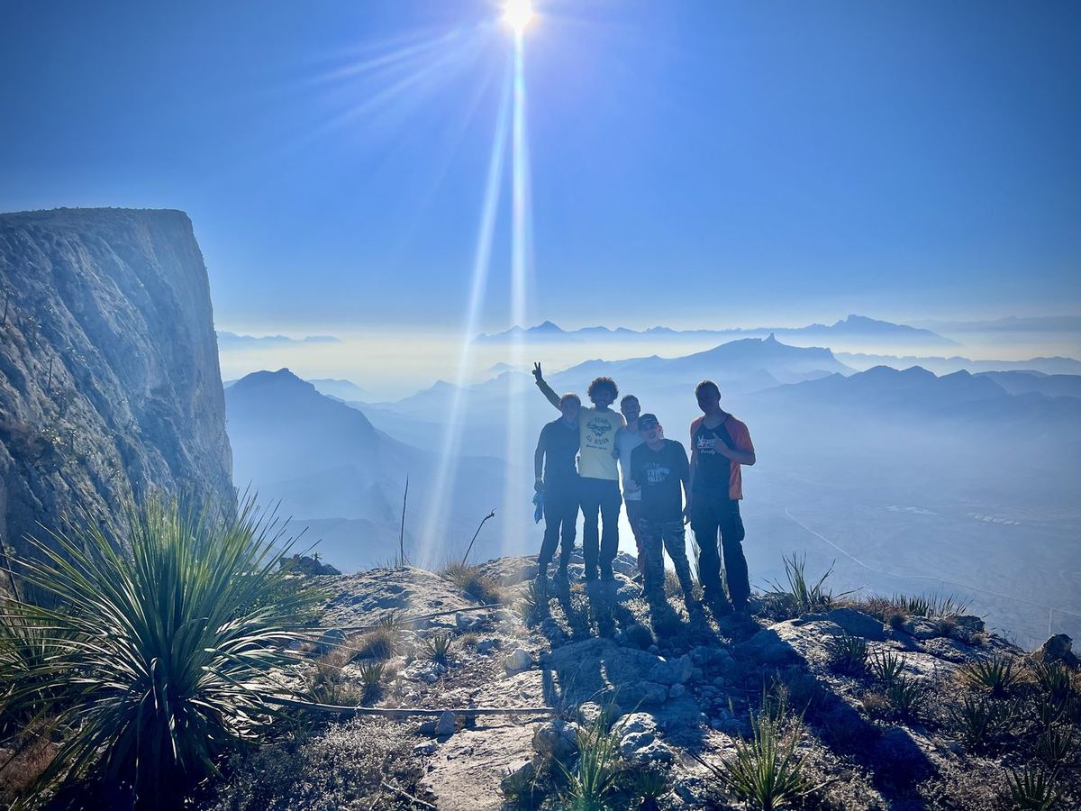 A group of friends on a mountain summit under a bright sunburst, above hazy ranges.