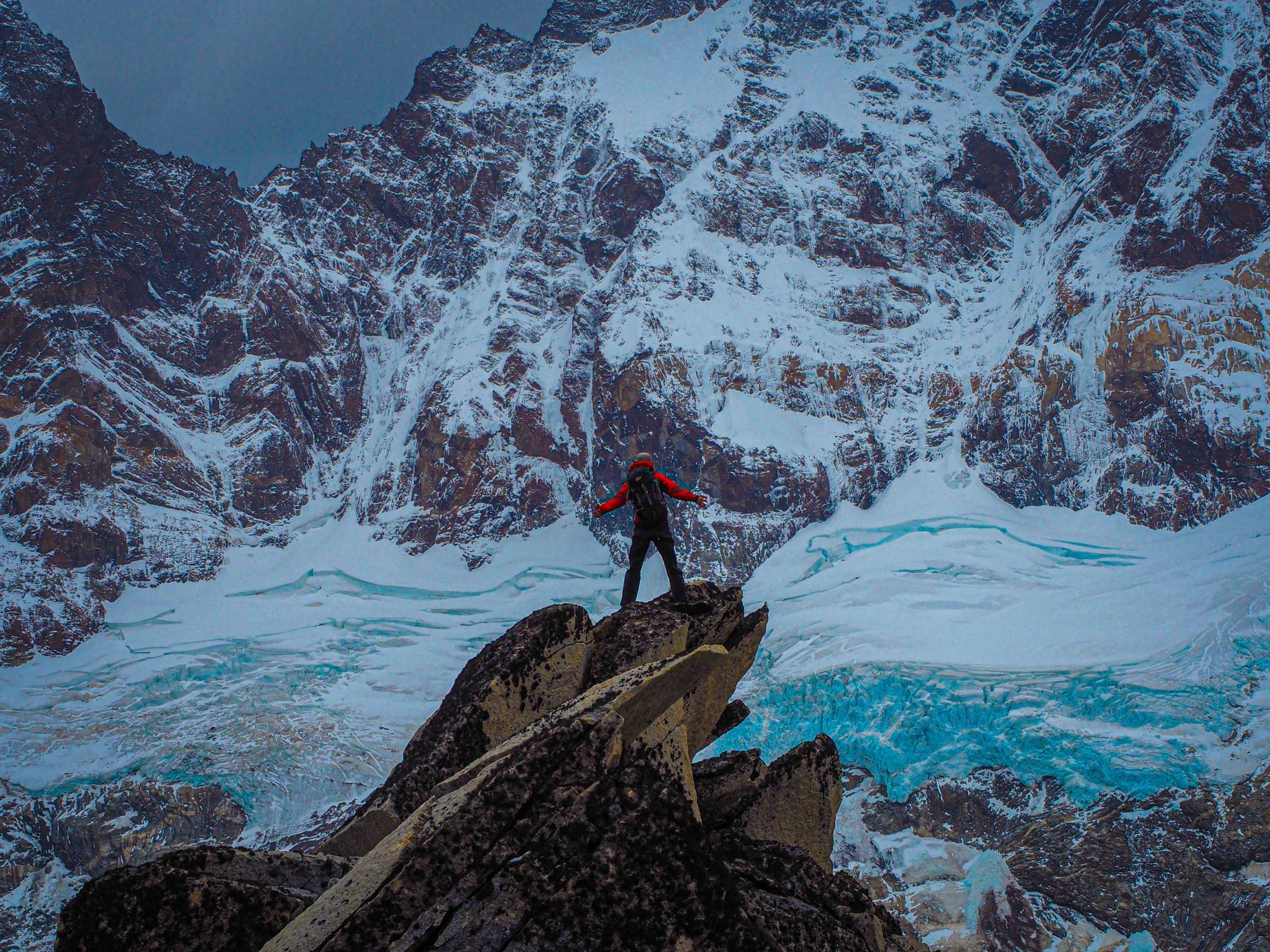 A lone figure on a rock spur below a vast wall of snow and glacier ice in Patagonia.